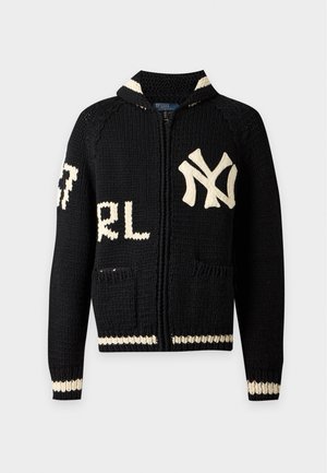Jeune homme debout portant un pull noir zippé avec des textes et logos blancs, un pantalon noir et des chaussures noires sur un fond clair uni.