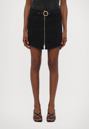 Jupe en denim noire avec une fermeture éclair sur le devant, une ceinture avec une boucle dorée ronde, et un ourlet évasé. Portée avec des sandales à talons noires à lanières.