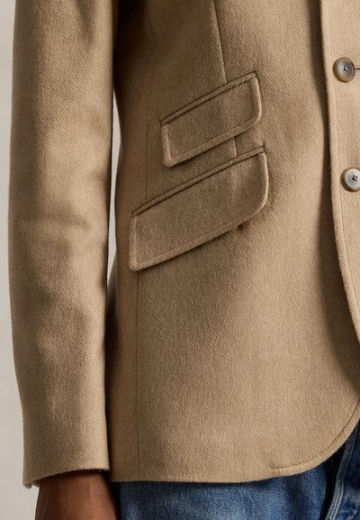 Smėlio spalvos vilnonis švarkas su žuvies kaulo raštu. Turi dvi užsegamas kišenes, vieną sagos uždarymą ir tekstūruotą audinį. Laisvas stilius su apvalia apačia.