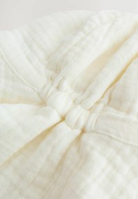 Tissu en coton couleur crème avec une surface texturée et matelassée. Présente un détail de nœud froncé au centre en haut. Doux et léger.