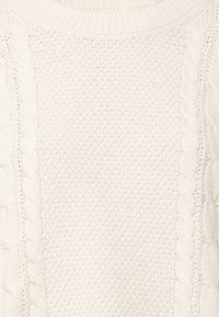 Pletený svetr krémové barvy s texturovaným copánkovým vzorem po stranách, žebrovaným výstřihem a měkkou, hustou pletenou strukturou.