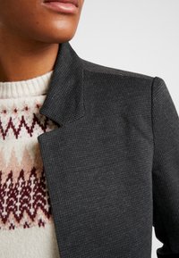 Veste texturée grise avec un col cranté, portée sur un pull crème présentant un motif géométrique rouge et marron. Vue en gros plan.