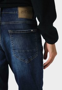 Tamsiai mėlynos džinsinės kelnės su tekstūruotu paviršiumi, juosimo juostoje yra rudos odos lopas, o nugaroje du kišenės su siuvimo detalėmis.