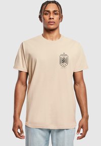 Tricou din bumbac bej, cu logo imprimat în negru în formă de scut, având cuvântul "ESPAÑA" și un design cu coroana, mâneci scurte și guler rotund.