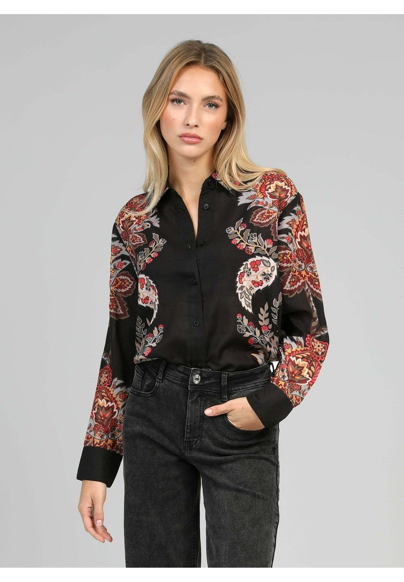 Chemise noire à boutons avec des motifs floraux en rouge, orange et gris. Manches longues avec des poignets noirs contrastés. Portée avec un jean en denim foncé.