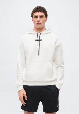 Jaunas vyras vilki baltą gobtuvą su juodomis raištelėmis ir juodus šortus, stovintis priešais paprastą baltą foną.