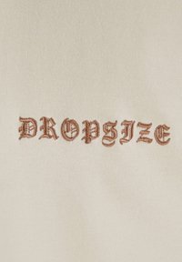 Cremefarvet stof med broderet tekst "DROPSIZE" i en stiliseret, brun skrifttype med indviklet detaljer. Glat tekstur.