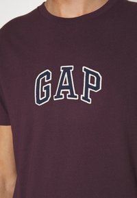 T-shirt en coton violet avec un logo "GAP" brodé en blanc et bleu marine sur la poitrine. Manches courtes et encolure ronde classique.