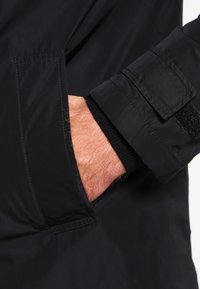 Černý zateplený rukáv bundy s hladkým vnějším povrchem, vybavený páskem na suchý zip a žebrovanými manžetami. Ruka viditelná v boční kapse.