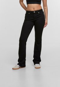 Pantalones de mezclilla negros acampanados con cintura de altura media, cinco bolsillos y detalles con botones plateados; textura suave y ajuste estructurado.