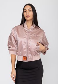 Donna che indossa una giacca bomber rosa chiaro lucida con chiusura frontale con cerniera e una gonna nera, posando con una mano nella tasca della giacca.