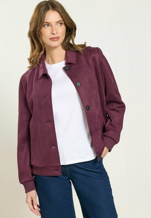 Donna con una giacca viola abbottonata sopra una camicia bianca e jeans blu scuro, in piedi con una mano nella tasca, che guarda a sinistra.