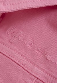 Pink cotton fabric with a textured finish and embroidered text reading "Life is." Visible stitching details along the seams.