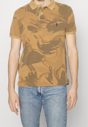 Mann trägt braunes Poloshirt mit Tarnmuster und blauem Logo auf der Brust sowie hellblaue Jeans, steht mit entspannten Armen.