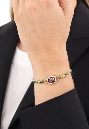 Armband aus Goldkette mit einem quadratischen lila Edelstein als Mittelpunkt, getragen am Handgelenk über einem schwarzen Ärmel und einem weißen Hemd.