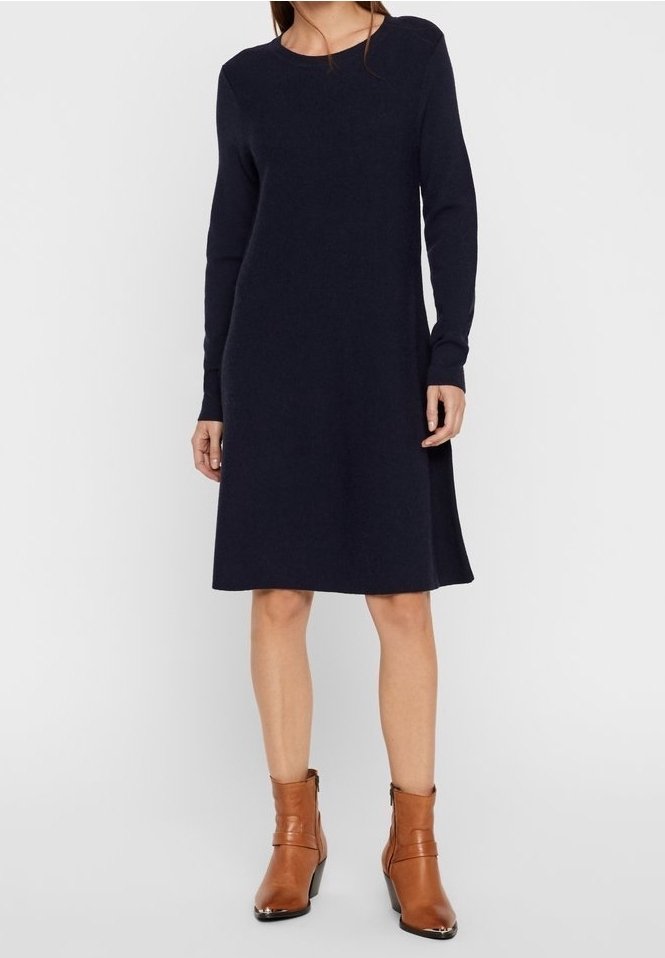 Femme portant une robe bleu marine foncé à manches longues et longueur genou, associée à des bottines en cuir marron à talons moyens, se tenant devant un fond uni.