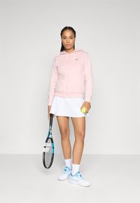 Sweat à zip rose clair avec poches avant, jupe de tennis blanche, tenant une balle de tennis et une raquette noire avec des accents bleus. Baskets et chaussettes blanches.