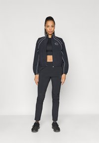 Chaqueta deportiva negra con detalles en blanco, puños elásticos y un diseño recortado, combinada con pantalones ajustados negros y zapatillas negras.
