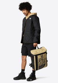 Svart quiltet jakke over en beige hettegenser, kombinert med svarte shorts. Bærer en to-farget tan og svart tote bag med logo. Svarte støvler.