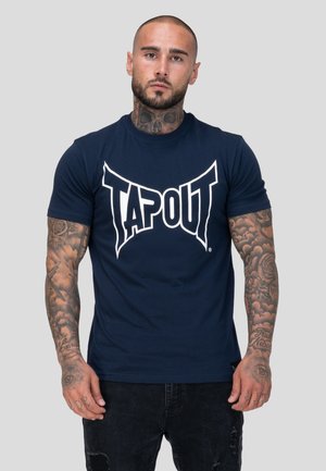 Man with tattoos on neck and arms wearing navy blue "TAPOUT" t-shirt and black ripped jeans, standing against a plain background.