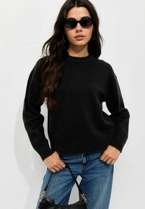 Mujer con suéter negro, vaqueros azules, gafas de sol negras y sosteniendo un pequeño bolso negro sobre un fondo liso y claro.