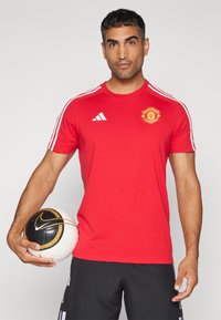Piros, rövid ujjú póló fehér csíkokkal, Manchester United logóval a mellkasán, fekete sorttal párosítva, és egy mintás focilabdát tartva.