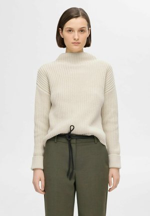 Beige knitted sweater with a high neck and ribbed texture, paired with dark green trousers featuring a black drawstring.