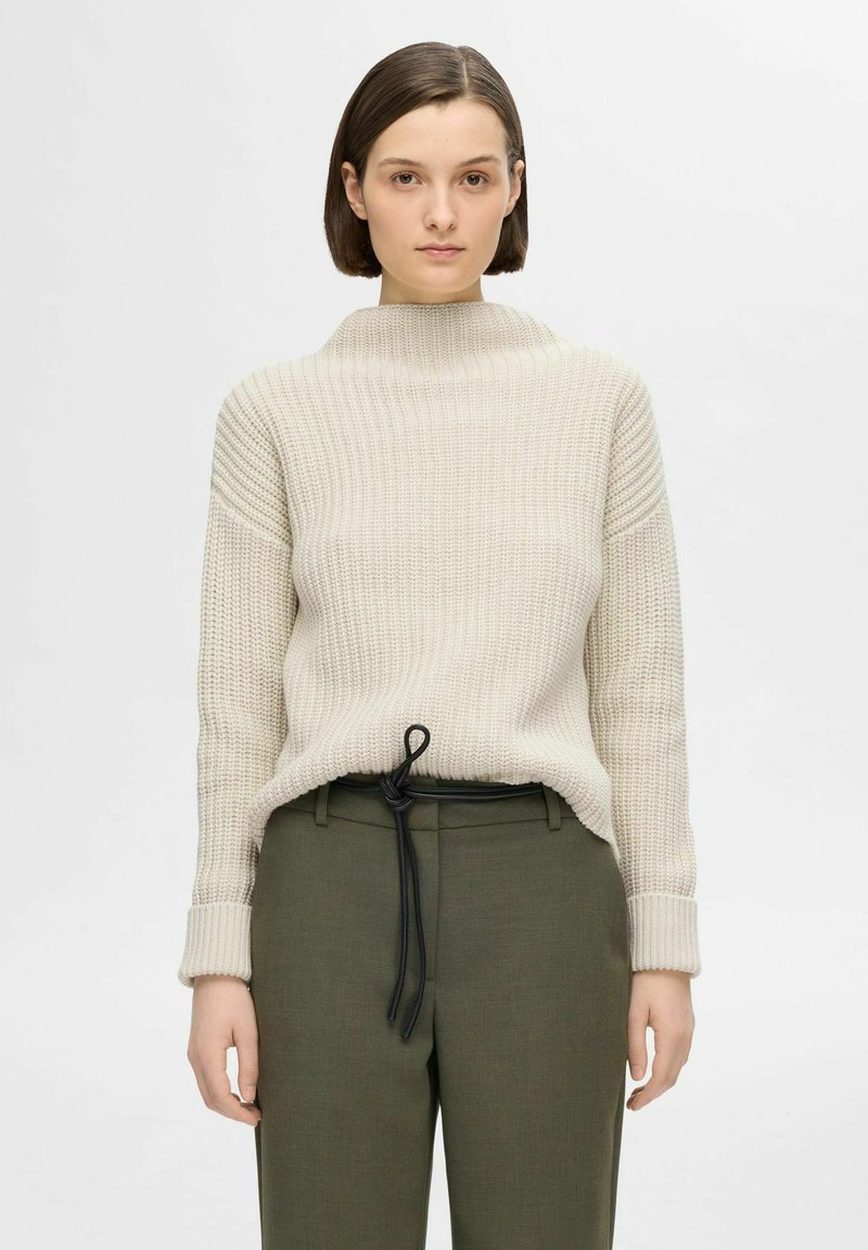 Pullover beige lavorato a maglia con colletto alto e texture a costine, abbinato a pantaloni verde scuro con cordino nero.