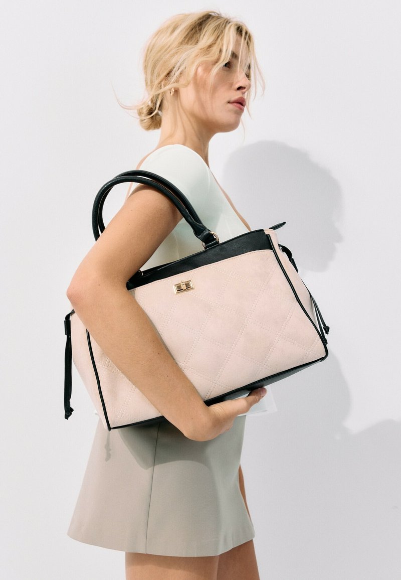Femme aux cheveux blonds tenant un grand sac à main rose clair et noir sur son épaule, portant un haut blanc sans manches et une jupe beige.