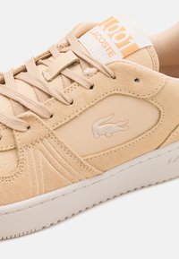 Baskets beige Lacoste avec panneaux en daim et cuir, lacets texturés, logo crocodile sur le côté, et étiquette de languette de marque.
