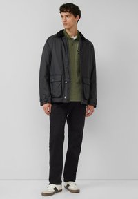 Czarna lekka kurtka z futrzanym kołnierzem, zapinana z przodu na guziki, z dwoma kieszeniami, noszona na zielonej koszuli, zestawiona z ciemnymi spodniami i sneakersami.