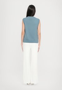 Ermeløs blå strikket topp med ribbestrikket kant, kombinert med hvite, vide, plissert bukser, sett fra baksiden. Glatt tekstur og enkel design.