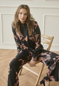 Vrouw met lang golvend haar zittend op een houten stoel, gekleed in een zwarte outfit met roze en beige bloemenpatronen, in een kamer met witte paneelwanden.