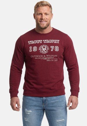 Blondyn z brodą w burgundowej bluzie z tekstem "Trout Trophy 1978" i jasnoniebieskich podartych dżinsach, stojący na białym tle.