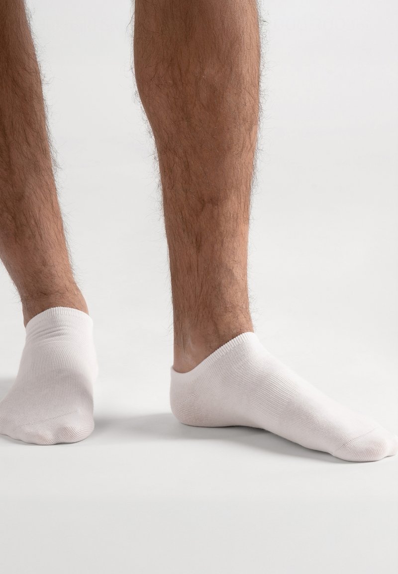 Weiße Knöchelsocken aus weichem Baumwollmaterial, mit geripptem Bündchen und figurbetontem Design, getragen auf einem nackten Fuß, teilweise sichtbare behaarte Beine.