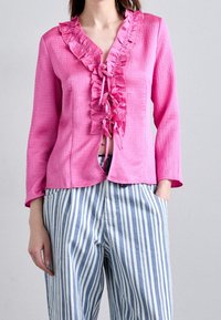 Blusa lunga a maniche con scollo a V arricciato e fiocco frontale di un vivace colore rosa, abbinata a pantaloni a righe blu e bianche. Dettaglio in tessuto testurizzato.