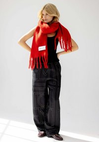 Rode wollen sjaal met franjerand, gecombineerd met zwarte, wijde broek met patroon en bruine schoenen. Label met de tekst "ARCTIC FOX."