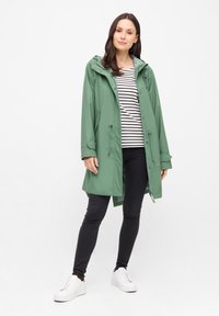 Grön vattentät jacka med huva, åtstramad midja och justerbara manschetter, bärs över en svart- och vitrandig topp, tillsammans med svarta byxor och vita sneakers.