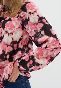 Donna che indossa una camicia a maniche lunghe con un motivo floreale rosa, nero e bianco, e con la mano nella tasca dei jeans di denim blu.
