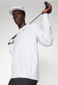 Bílý dámský golfový svetr s dlouhým rukávem a kulatým výstřihem, s malým černým logem. Bílá kšiltovka a golfová hůl jsou drženy přes rameno.