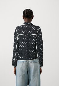 Quilted svart jakke med hvite prikkar, kontrastert av kvite kantar. Har ein tilpassa krage og slim fit-design, kombinert med lyse blå jeans.