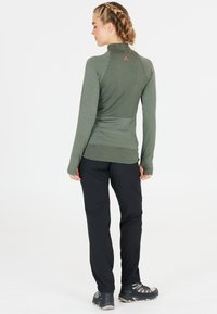 Langärmliges, olivgrünes Oberteil mit hohem Kragen und strukturiertem Stoff, kombiniert mit schwarzen Hosen und Schnür-Wanderschuhen.