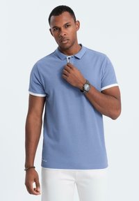 Světle modré polo tričko s krátkými rukávy a bílým lemem, které má knoflíkovou légu a vyšívaný detail blízko lemu.