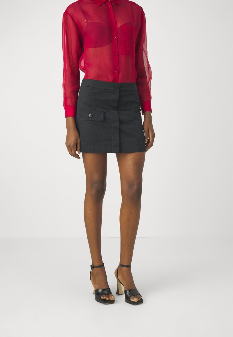 Chemise rouge transparente à manches longues avec fermeture à boutons, associée à une mini-jupe noire dotée de deux poches latérales. Sandales à talons hauts noires.