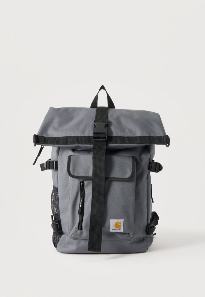 Zaino Carhartt grigio con chiusura a rotolo, cinturino nero con fibbia, tasca frontale con zip, tasca laterale in rete e toppa con logo del marchio sulla tasca frontale.