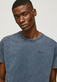 Slate blue cotton t-shirt with a crew neck and short sleeves. Features a small black “Pepe Jeans” logo on the left chest. Smooth texture.