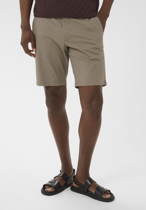 Mężczyzna ubrany w beżowe szorty do kolan i czarne sandały z odkrytymi palcami, stojący z jedną ręką w kieszeni na jednolitym tle.