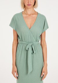 Vestido verde envuelto hecho de tela arrugada con mangas cortas y un profundo escote en V. Presenta un cinturón atado en la cintura para un detalle adicional.