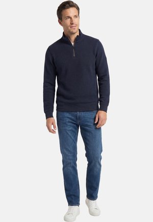 Homme portant un pull marine à col zippé, un jean bleu et des baskets blanches, debout devant un fond clair uni.