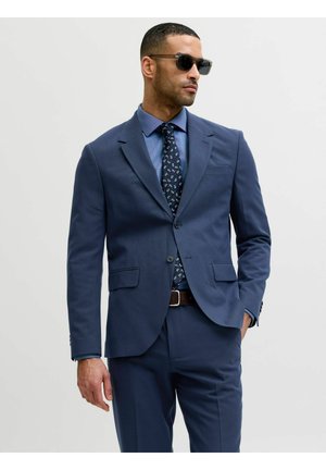 Man draagt een marineblauw pak, blauw overhemd, een stropdas met patroon, een bruine riem en zwarte zonnebril, staand met één hand in zijn zak tegen een lichte achtergrond.